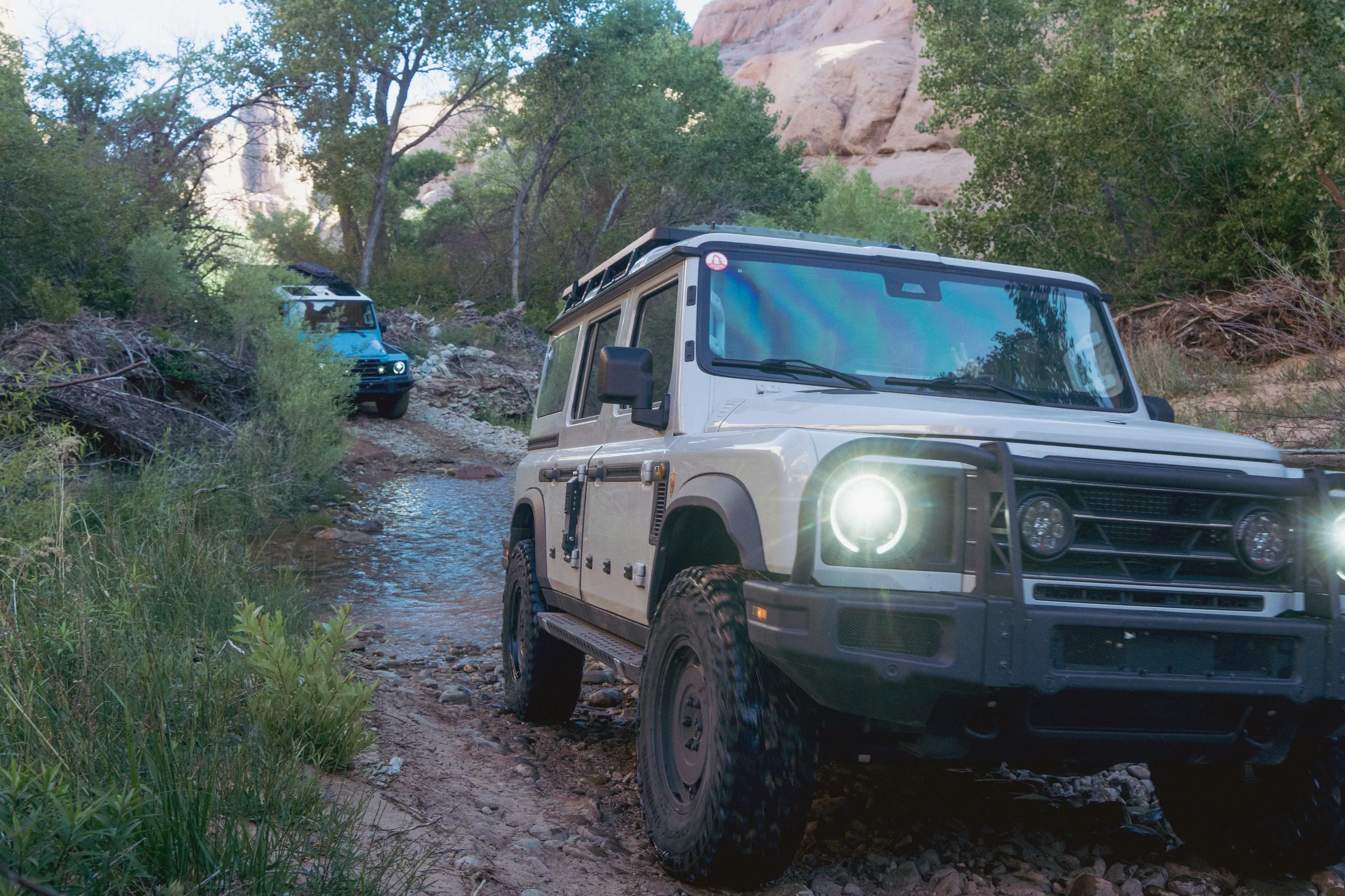 A white Grenadier with headlights on in a green canyon wash